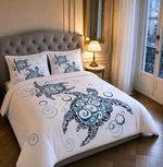 A bed dressed in a white duvet cover and matching pillowcases featuring a large dark teal turtle illustration with swirling patterns and smaller light blue bubble motifs, styled in a bedroom with a tufted gray headboard, bedside lamp, mirror, and window.